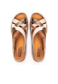 Sandalias tipo slide con diseño de tiras cruzadas en beige, crema y dorado metálico. Parte superior de cuero, plantilla marrón y detalles de suela trenzada. Tacón plano.