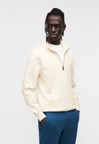 Εκρού φούτερ με λαιμόκοψη και φερμουάρ, με μπροστινή τσέπη, φορεμένο πάνω από λευκό πουκάμισο, συνδυασμένο με μπλε παντελόνι. Απαλή υφή, λιτός σχεδιασμός με ελάχιστες λεπτομέρειες.