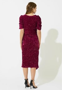 Femme portant une robe à sequins violette foncée aux genoux avec des manches courtes bouffantes, associée à des sandales noires à talons hauts, se tenant de dos.