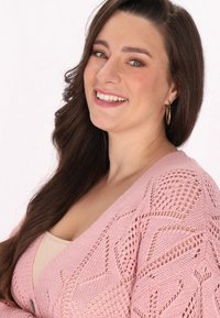 Világos rózsaszín kötött kardigán, nyitott szemmintával, mély V nyakkivágással és hosszú ujjal. Az anyag lágy, textúrált megjelenésű.