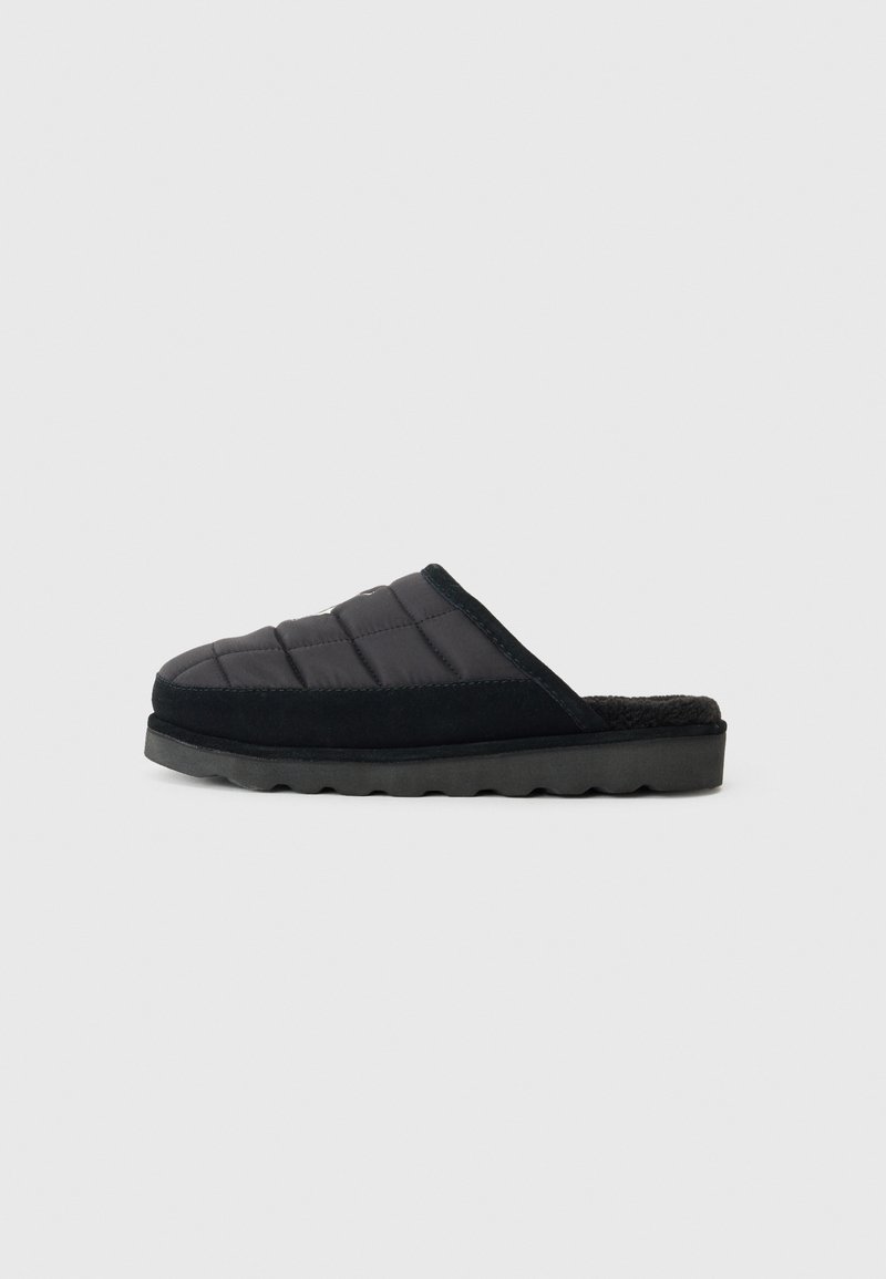 Zwarte instapslipper met een bovenkant van gequilt textiel, zwarte suede accenten, een zachte bontvoering en een rubberen zool met een golfpatroon.