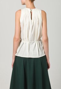 Vit ärmlös blus med veckad detalj på ryggen, nyckelhålsöppning som stängs med knapp, kombinerad med en grön kjol. Mjuk tygstruktur.