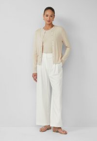 Beiger Cardigan mit Knopfverschluss, kombiniert mit einem beigen Oberteil und weißen, weit geschnittenen Hosen. Die Schuhe sind flache Sandalen mit Knöchelriemen.