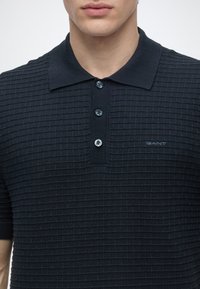 Gros plan d'une personne portant un polo noir texturé avec trois boutons et un petit logo GANT sur la poitrine.