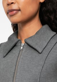 Γκρι φερμουάρ μπουφάν με μεγάλο γιακά, με λεία υφή και μεταλλική λεπτομέρεια στο φερμουάρ, τοποθετημένο στο λαιμό.