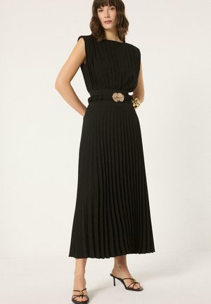 Γυναίκα στέκεται φορώντας ένα μακρύ μαύρο πλισέ φόρεμα χωρίς μανίκια με φλοράλ αγκράφα στη ζώνη και μαύρα σανδάλια με λουράκια και τακούνια.