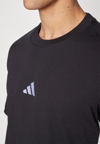 Svart bomullströja med rund halsringning och korta ärmar, med en liten, ljusblå tre-stripes-logotyp på vänster bröst.