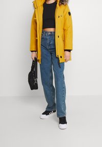 Parka amarilla con capucha forrada de piel, top corto negro, jeans azul de cintura alta y zapatillas negras. También se incluye un bolso negro.