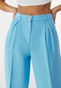 Light blue trousers featuring front pleats, a button closure, and a smooth fabric texture. Tailored fit with side pockets.