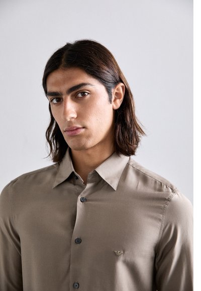 Cămașă button-up maro deschis, cu mâneci lungi, având o textură netedă, nasturi negri și un logo subtil pe piept.