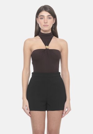 Donna che indossa un top nero senza maniche con chiusura dorata e pantaloncini neri a vita alta, in piedi davanti a uno sfondo chiaro e uniforme.