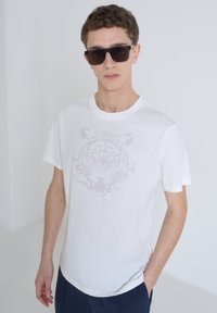 Camiseta de algodón blanca con un gráfico de un tigre gris claro en el frente. La camiseta tiene mangas cortas y un escote redondo.