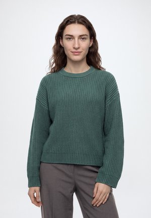Jeune femme aux cheveux bruns ondulés porte un pull vert tricoté et un pantalon gris, debout devant un fond blanc uni.