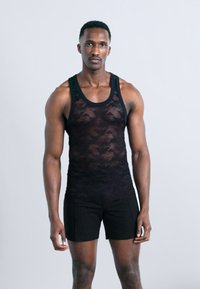 Αθλητικός άνδρας που φοράει μαύρο αμάνικο τοπ με σχέδια και μαύρο σορτς, στέκεται μπροστά σε απλό φωτεινό φόντο.