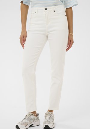 Person trägt weiße Jeans mit hohem Bund, gestreiftes Oberteil und weiße-graue New Balance Sneakers, steht vor einfarbigem Hintergrund.