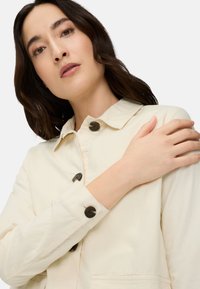 Woman with brown hair wearing a cream-colored button-up jacket, holding her left shoulder with her right hand against a plain background.