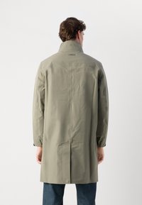 Gabán largo de color verde oliva claro, fabricado con material impermeable, que presenta un cuello alto, puños elásticos y un detalle de costura central en la parte posterior.