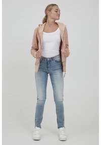 Felpa con zip di colore rosa chiaro con motivo testurizzato, canotta bianca e jeans skinny blu sbiaditi, abbinati a sneakers bianche.
