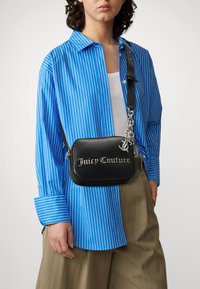 Černá texturovaná krosbody taška se stříbrným logem "Juicy Couture", s řetízkovým popruhem a kompaktním obdélníkovým tvarem, nošena přes modrou pruhovanou košili.