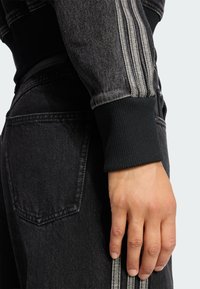 Crna traper jakna s sivim rebrastim manšetama i kontrastnim bočnim prugama. Tkanina je teksturirana, s vidljivim šavovima i detaljima džepova.