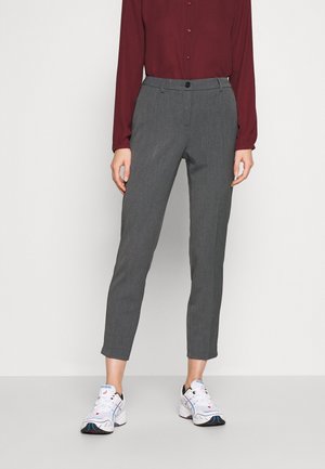 Femme portant un pantalon gris slim longueur cheville, une chemise bordeaux à manches longues boutonnée, et des chaussures de sport blanches, debout devant un fond uni.