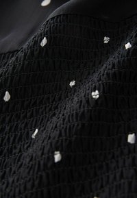 Svart tyg med texturerad smocking och vita blommotiv broderi accenter. Materialet verkar mjukt och lätt med en avslappnad design.