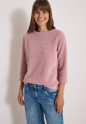 Femme souriante aux cheveux bruns lisses portant un haut rose à manches longues et un jean bleu, debout devant un fond clair uni.