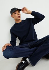 Jūras spēku adīts džemperis ar faktūru, tumši džinsi, melna cepure un melni kedas ar baltām akcentiem. Rokas novietotas izsistā pozā.