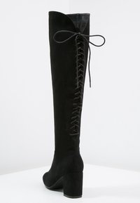 Stivali oltre il ginocchio in suede nero con tacco a blocco, caratterizzati da una stringa posteriore regolabile per una vestibilità personalizzata. Texture liscia complessiva.