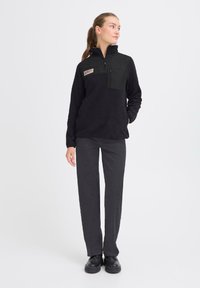 Schwarzer Fleece-Pullover mit Viertelreißverschluss, kontrastierender Einsätze und Brusttasche. Trägt dazu schwarze gerade Hosen und schwarze Schuhe.