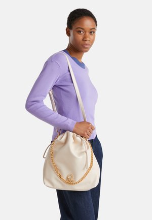 Sac en cuir crème avec un système de fermeture à cordon, bandoulière en chaîne dorée et accent de logo subtil. Porté avec un pull violet clair et un jean foncé.