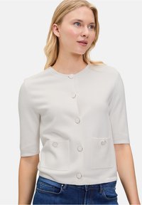 Modelka z włosami do ramion w kolorze blond nosi jasnobeżową bluzkę z srebrnymi guzikami i kieszeniami, w połączeniu z niebieskimi dżinsami, delikatnie się uśmiechając.
