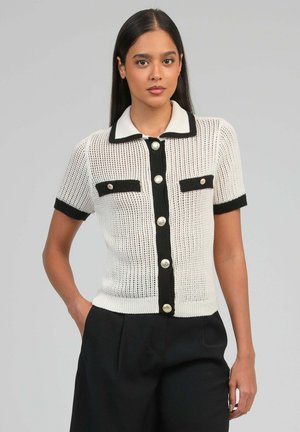 Femme portant un top en maille blanc à manches courtes avec bordure noire, boutons dorés et deux poches avant, associé à un pantalon noir.