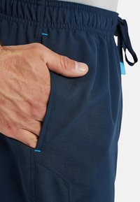 Pantalones deportivos de color marino con cintura elástica, con una mano en el bolsillo lateral y un acento azul en la costura del bolsillo.