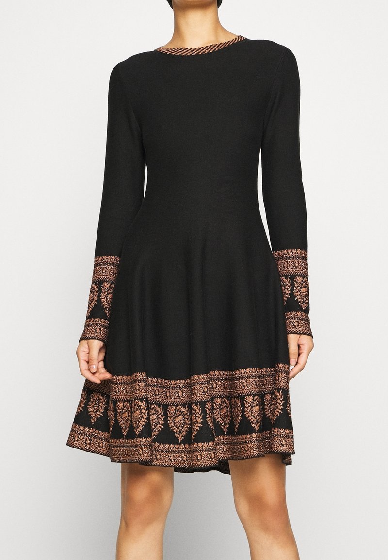 Robe noire à manches longues avec broderie florale cuivrée sur les poignets, l'ourlet et le décolleté, portée par une personne debout devant un fond clair.