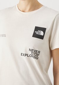 Camiseta de algodón color crema con un logotipo cuadrado negro que incluye "THE NORTH FACE" y la frase "NEVER STOP EXPLORING" en texto negro en negrita.