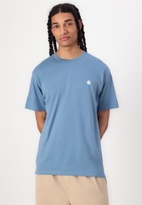 T-shirt en coton bleu clair à manches courtes, col rond, avec un petit logo blanc sur la poitrine gauche. Texture lisse, coupe décontractée.