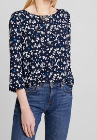 Marineblaue Bluse mit floralen Mustern in Weiß und Blau, runder Ausschnitt, Dreiviertelärmel, leichtes Material, kombiniert mit blauen Jeans.