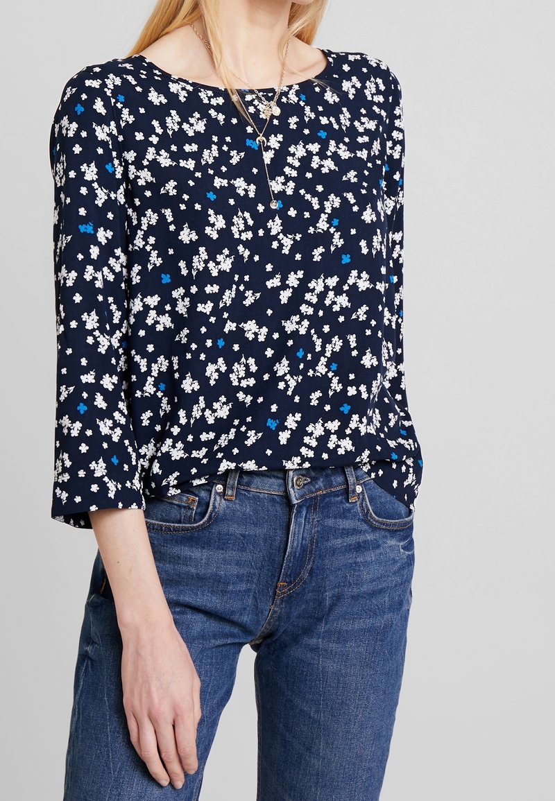Marineblaue Bluse mit floralen Mustern in Weiß und Blau, runder Ausschnitt, Dreiviertelärmel, leichtes Material, kombiniert mit blauen Jeans.