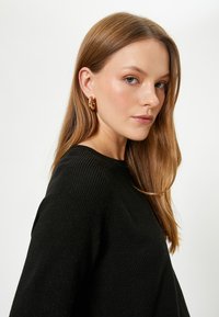 Maglione nero a coste con scollatura tonda, dotato di maniche lunghe. La modella indossa orecchini a cerchio dorati e ha i capelli lisci, di lunghezza spalle.