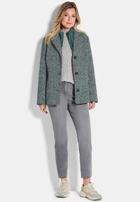 Femme portant un pull en tricot torsadé gris, une veste en chevrons verte, un jean gris et des baskets beiges sur un fond uni.