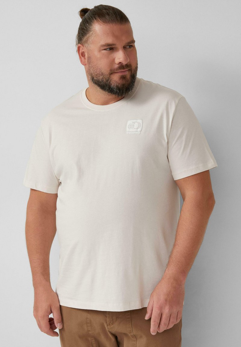 Helles beiges T-Shirt mit kurzen Ärmeln aus weicher Baumwolle und einem kleinen weißen Logo-Patch auf der linken oberen Brust. Glatte Textur, normale Passform.