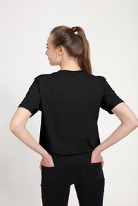 Camiseta negra de algodón, mangas cortas, cuello redondo, textura suave; combinada con vaqueros negros, manos en los bolsillos, vista desde atrás.