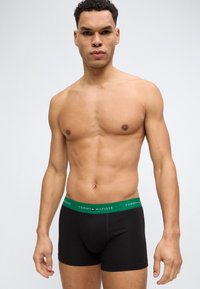 Άνδρας χωρίς πουκάμισο, φοράει μαύρα boxer με πράσινη μέση Tommy Hilfiger, στέκεται μπροστά σε απλό φωτεινό φόντο.