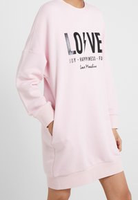 Person som bär en ljusrosa långärmad sweatshirtklänning med svart texten "LOVE" och orden "JOY HAPPINESS FUN" på framsidan.
