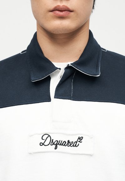 Κοντινό πλάνο ενός ατόμου που φοράει πουκάμισο με γιακά σε μαύρο και άσπρο, με κεντημένο το λογότυπο "Dsquared²" στο στήθος.