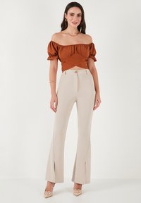 Off-shoulder top i rustfarvet med rynkedetaljer og puffærmer; beige flared bukser med slitser; parret med lyse farvede sko.