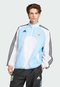 Νεαρός άνδρας που φοράει ελαφρύ μπλε και λευκό μπουφάν ποδοσφαίρου Adidas Argentina με μαύρο παντελόνι Adidas, σε απλό φόντο.