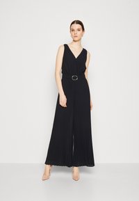 Mono negro con pliegues, escote en V, cintura ceñida con un cinturón de hebilla redonda y piernas anchas. La modelo lleva tacones nude y accesorios mínimos.
