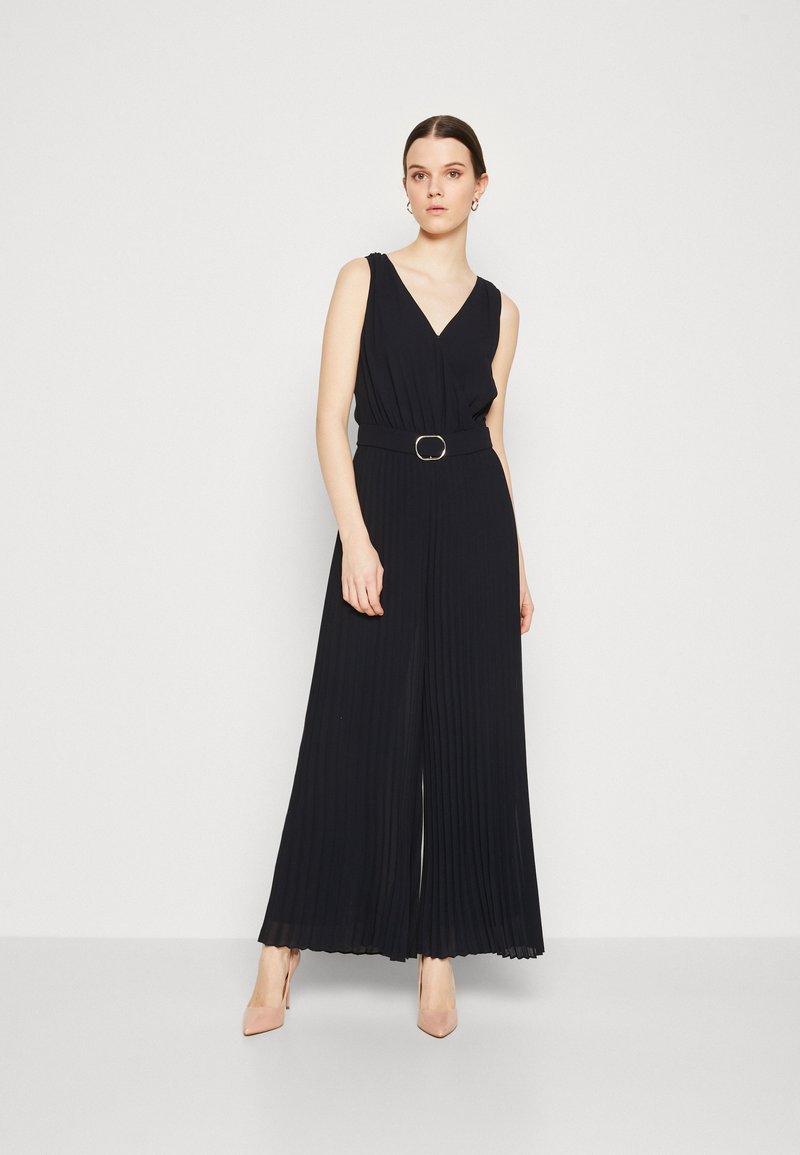 Mono negro con pliegues, escote en V, cintura ceñida con un cinturón de hebilla redonda y piernas anchas. La modelo lleva tacones nude y accesorios mínimos.